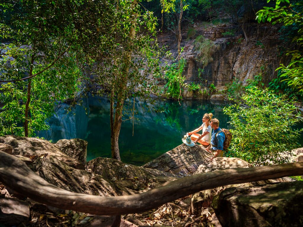 A topaz blue waterhole at Emma Gorge in El Questro, surrounded by ochre rock formations and Australian native trees, with a man and woman sitting on the edge, admiring the serene Kimberley landscape.