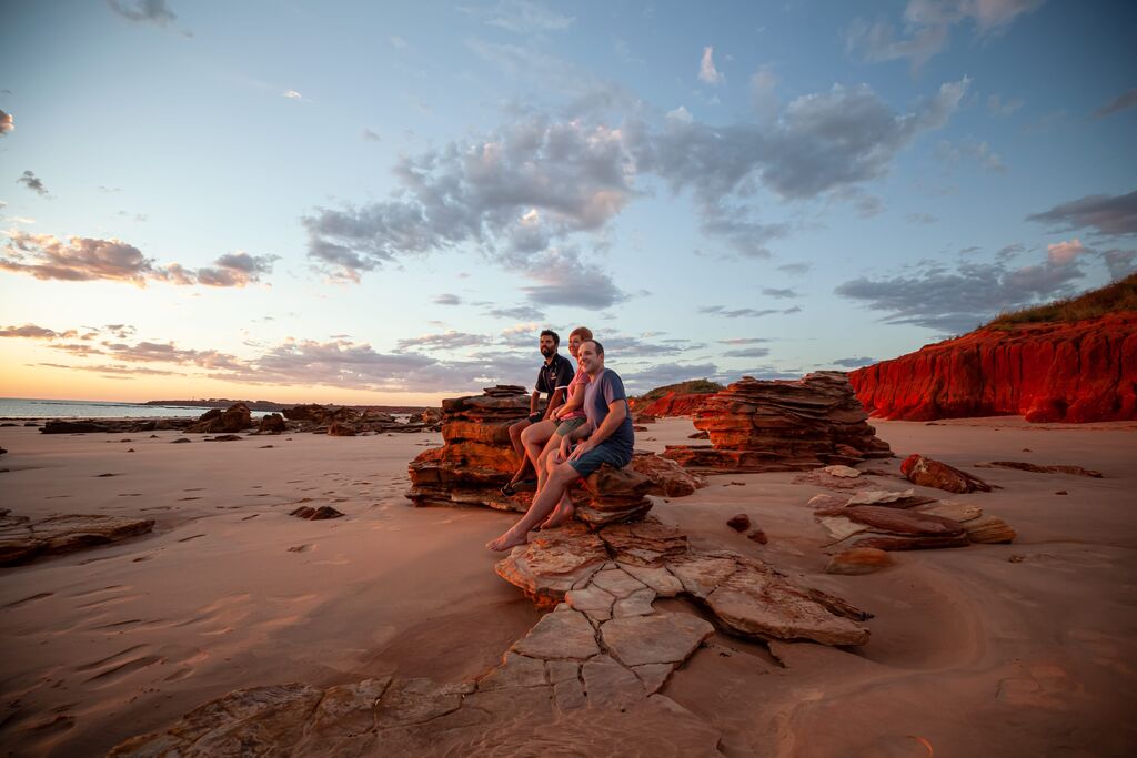 Three people sitting on ochre-colored rocks, gazing out at the sea during a serene sunset in the Kimberley region near Broome, Western Australia, with a few clouds scattered across the blue sky.